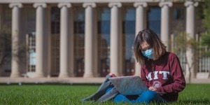 A student in a mask and MIT sweatshirt works on her laptop on the grass in Killian Court