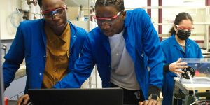 Two young men in blue coats and goggles look at a laptop together. A woman is in the background
