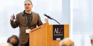 Professor Aleksander Mądry gestures with open hands behind a podium labeled ALC. He wears a brown jacket and an ALC badge.