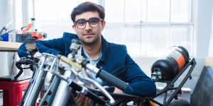 Adi Mehrotra is shown in a workshop, leaning on the handles of what appears to be a motorcyle. Large windows and a workbench appear in the background.