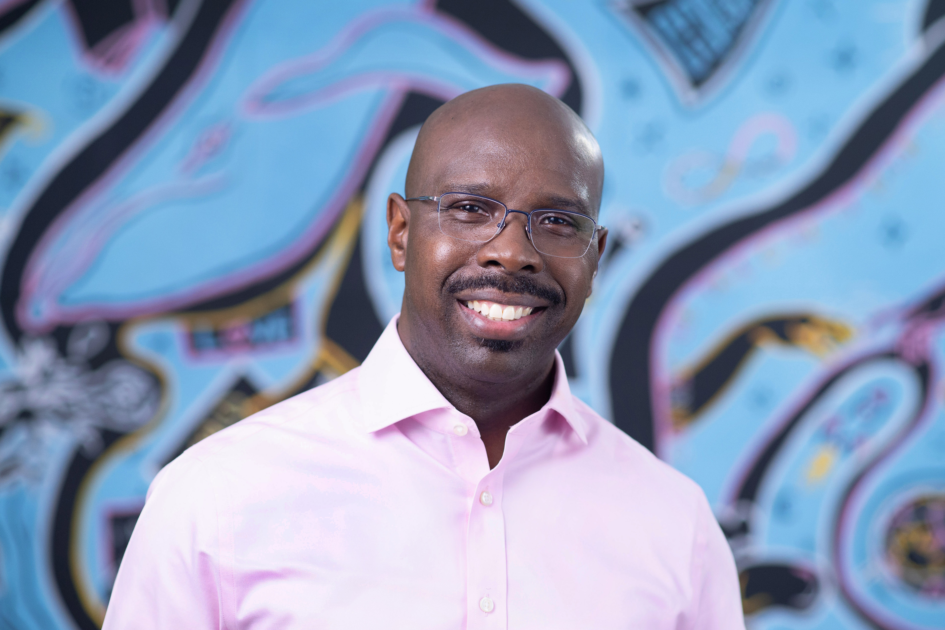 Portrait of Michael G. Johnson wearing a pink shirt and standing in front of a blue background with black swirls.