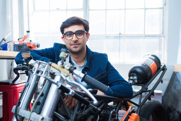 Adi Mehrotra is shown in a workshop, leaning on the handles of what appears to be a motorcyle. Large windows and a workbench appear in the background.