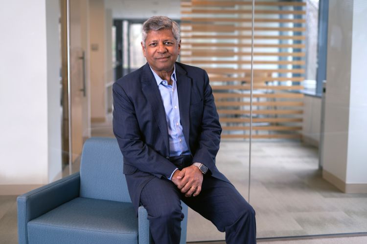 Sudhakar Kesavan wears a blue suit and sits casually on the arm of a blue armchair. The background suggests an office setting.