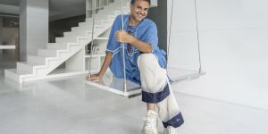 Parmesh Shahani sits on a swing in a spare white room with white steps in the background. He wears a blue shirt and white pants.