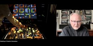 A split screen image shows, on the left, an arm reaching toward a set of blocks arrayed on a table. A screen in the background shows blocks in different colors. On the right is Kent Larson speaking with bookcases in the background.
