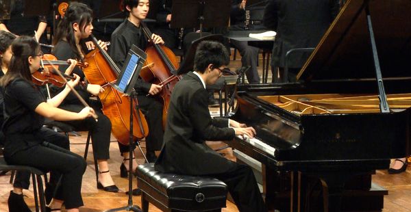 A pianist in a black suit sits on a bench playing a grand piano. Members of an orchestra appear behind him, notably two cellists and two violinists. Black music stands obscure other people.
