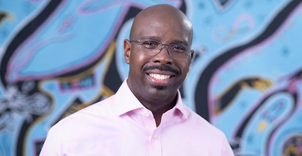Portrait of Michael G. Johnson wearing a pink shirt and standing in front of a blue background with black swirls.