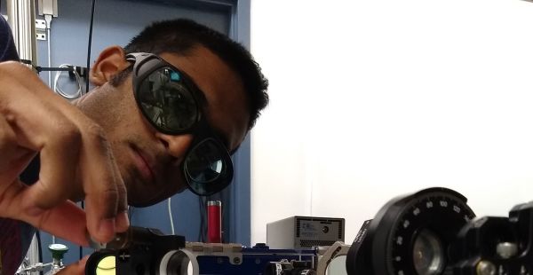 Sahil Pontula is shown in dark glasses leaning over some equipment that includes an array of lenses and various metal parts. He appears to be adjusting some element of the machinery.