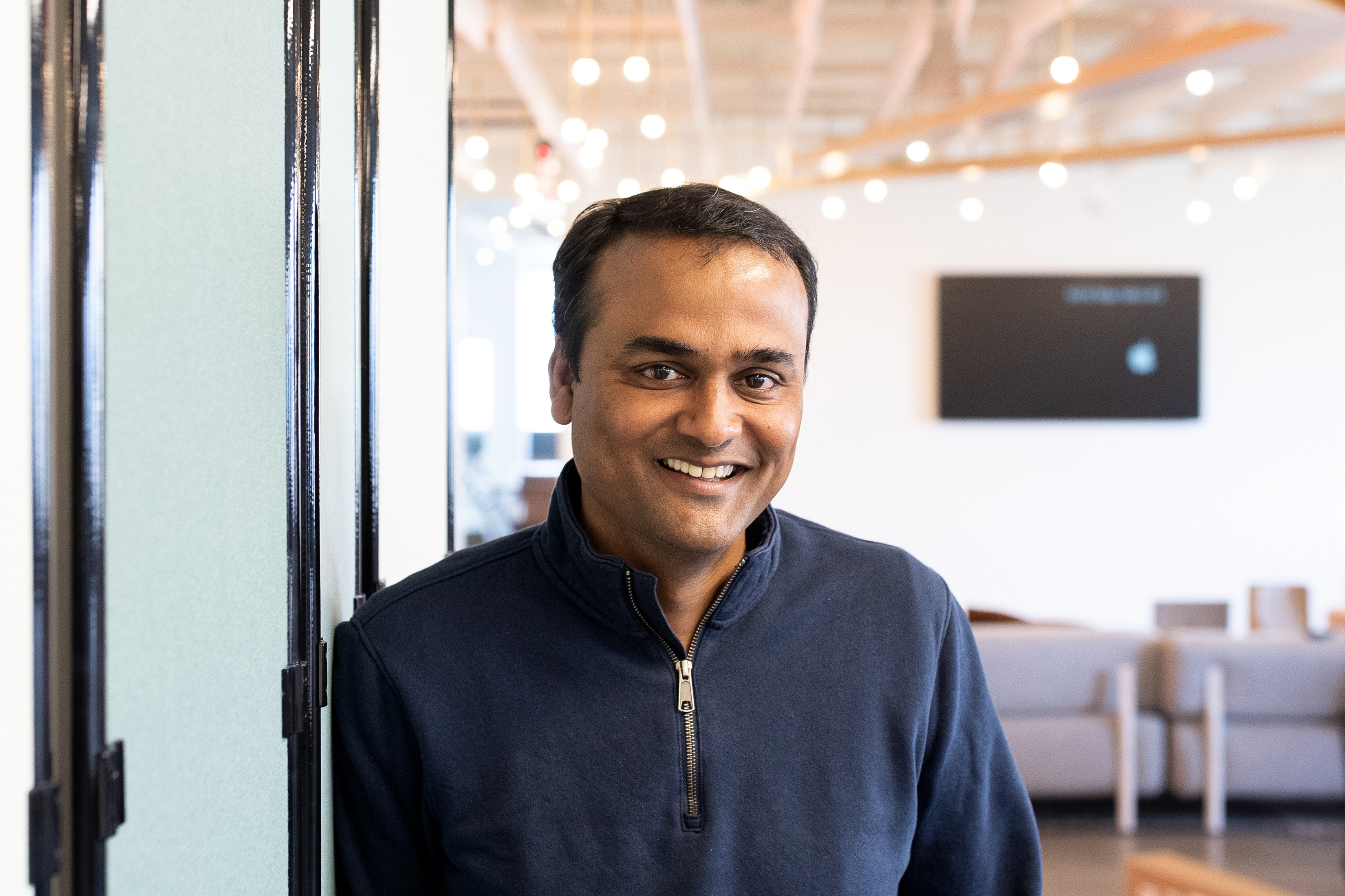 A photo of Ravi Inukonda standing inside leading against a class wall with a TV and couch behind him