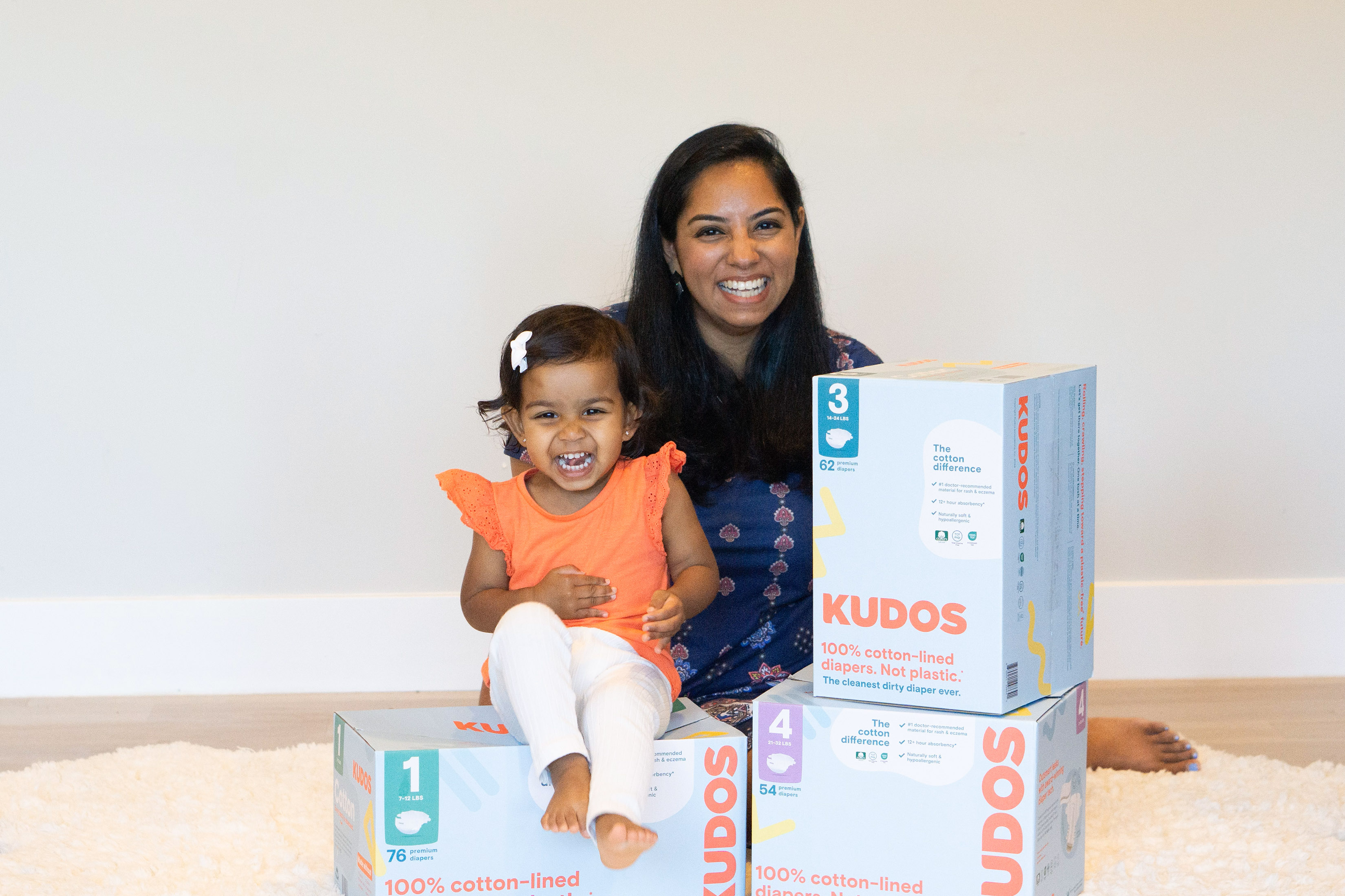 A photo of Amrita Saigal in a white room sitting on a white rug with boxes of Kudos diapers in front of her and a child sitting on one of the boxes