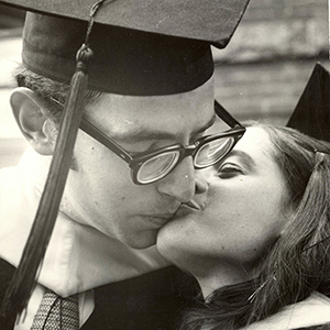 A black and white photo of a man in a cap and gown kissing a woman