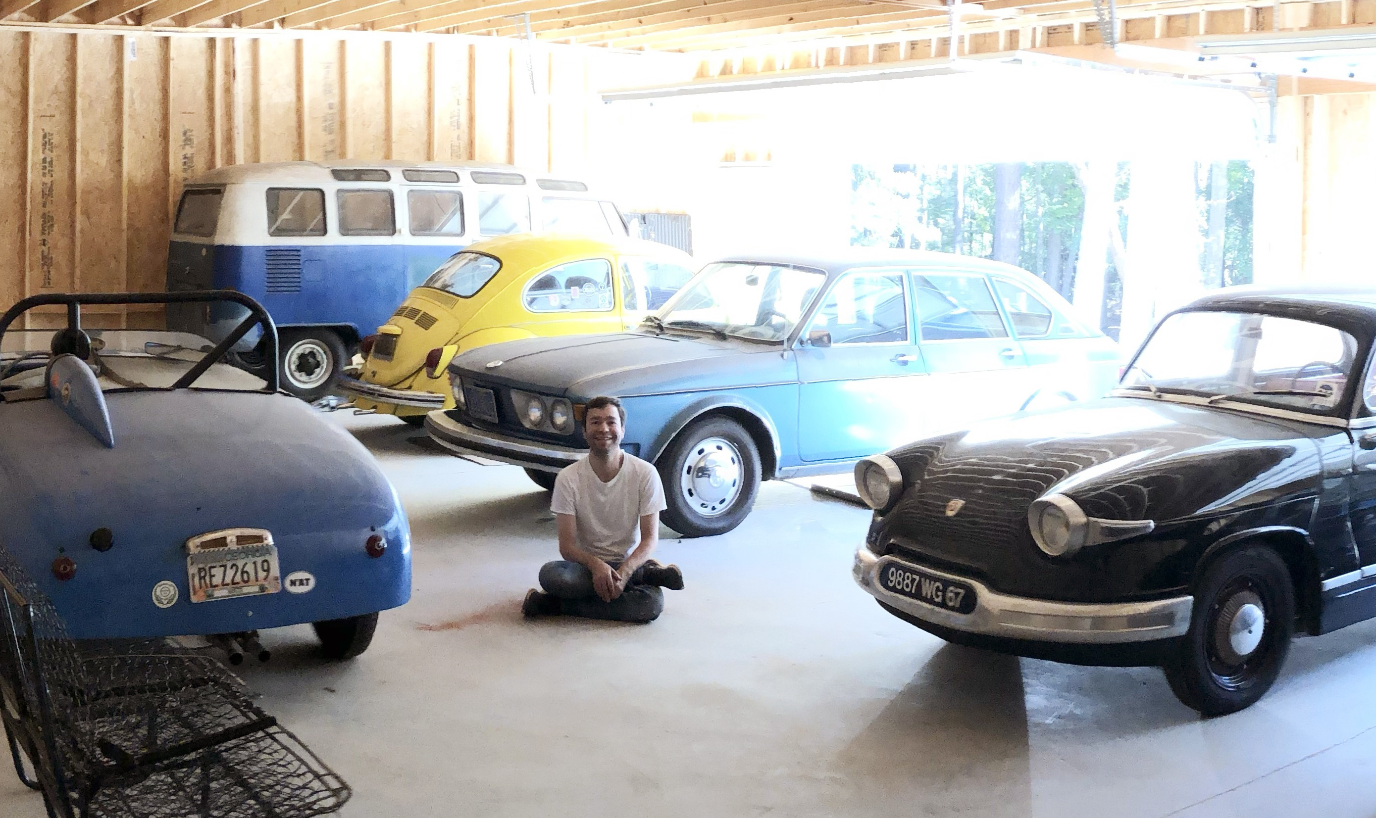A photo of David Lucsko sitting on the floor of a garage surrounded by old cars 