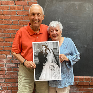 Photo of Jo E. Comparato and Thomas Frank Comparato ’56 standing in front of a wall with brick on the left and chalkboard on the right, holding a black and white image wedding photo