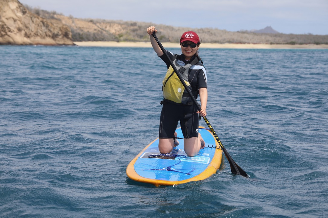 Woman wearing a wetsuit kneeling on a paddleboard in the ocean with a beach and rocky coastline behind her