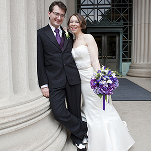 A photo of Adam Ganderson and Rebecca Richkus in a wedding dress and suit leaning against a column with a building entrance behind