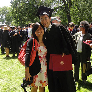 A photo of Kathy Li and Chris Su who is holding a diploma and wearing a cap and gown outside in the grass