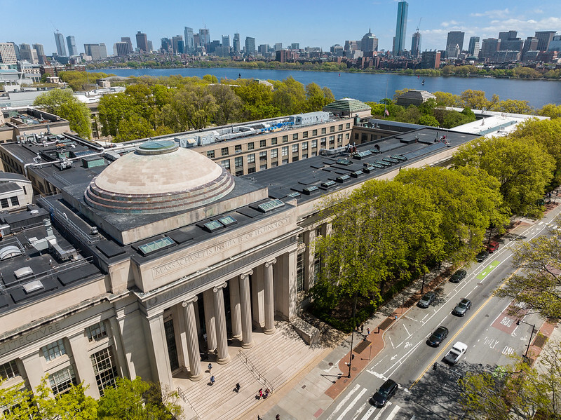 Aerial photo of MIT's campus from 77 Mass Ave with building tops and trees in the forefront and the Charles River and Boston city skyline behind