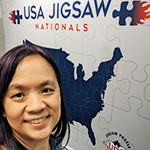 A photo of MIT alum Tammy McLeod standing in front of a banner that says USA Jigsaw Nationals