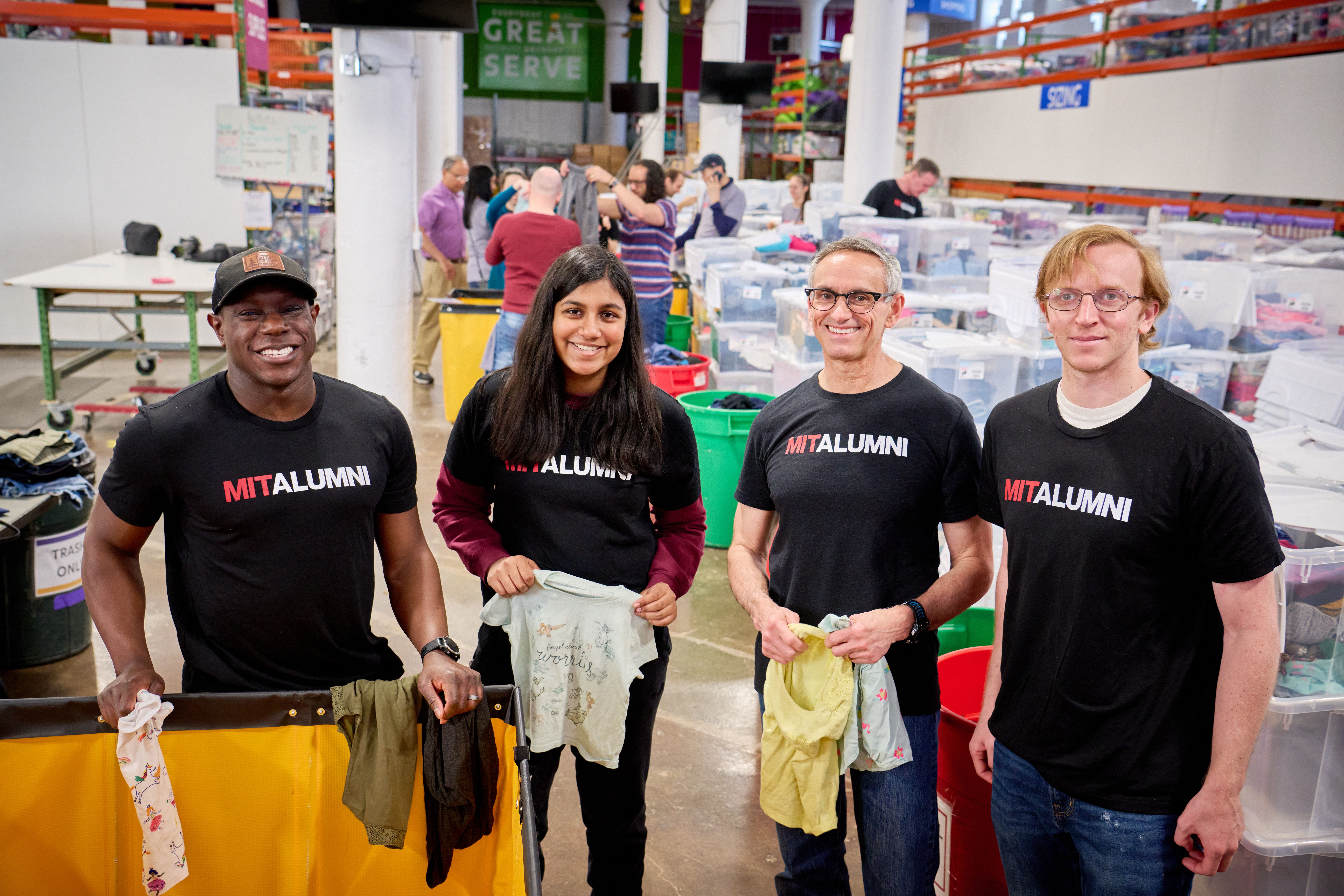 Four MIT Alumni wearing black shirts with MIT Alumni on the front stand in a warehouse holding clothes