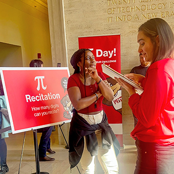 A woman holds her hand to her chin and stares upward as another woman looks at a notebook and points at it. At left is a big poster with the symbol for pi and the words Recitation, How many digits can you recite? A poster in the background shows Tim the Beaver and the words Day! and challenge.