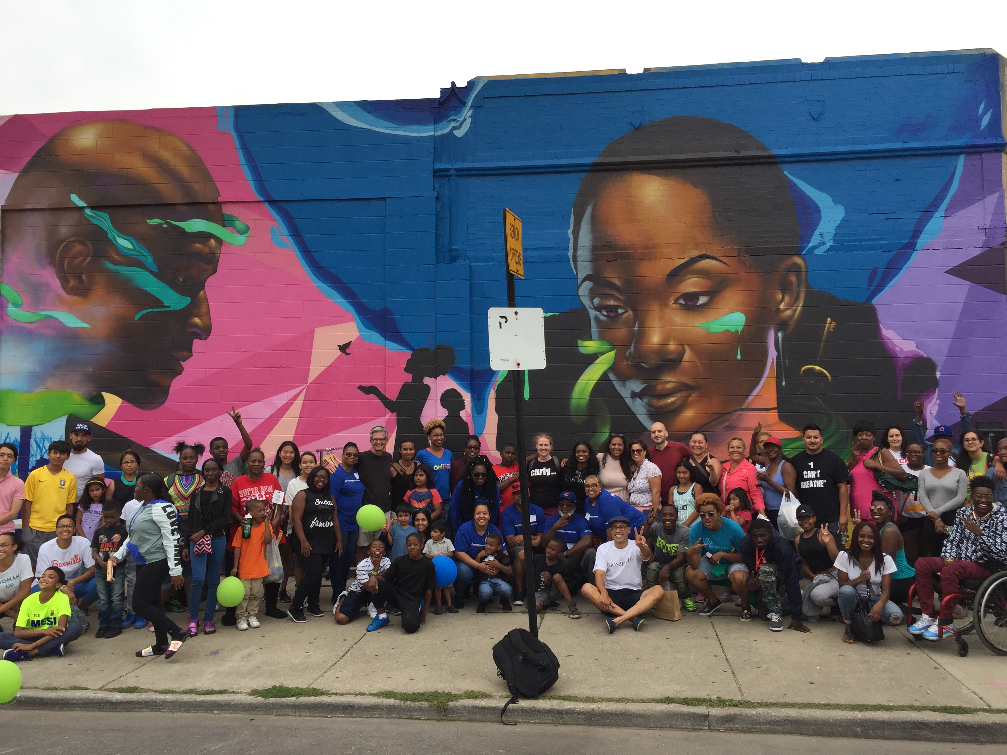 A large group on a sidewalk in front of a mural of a man and woman's faces