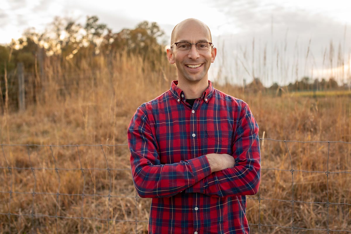 Smiling man with glasses, no hair, and plaid shirt standing in a field with arms crossed 