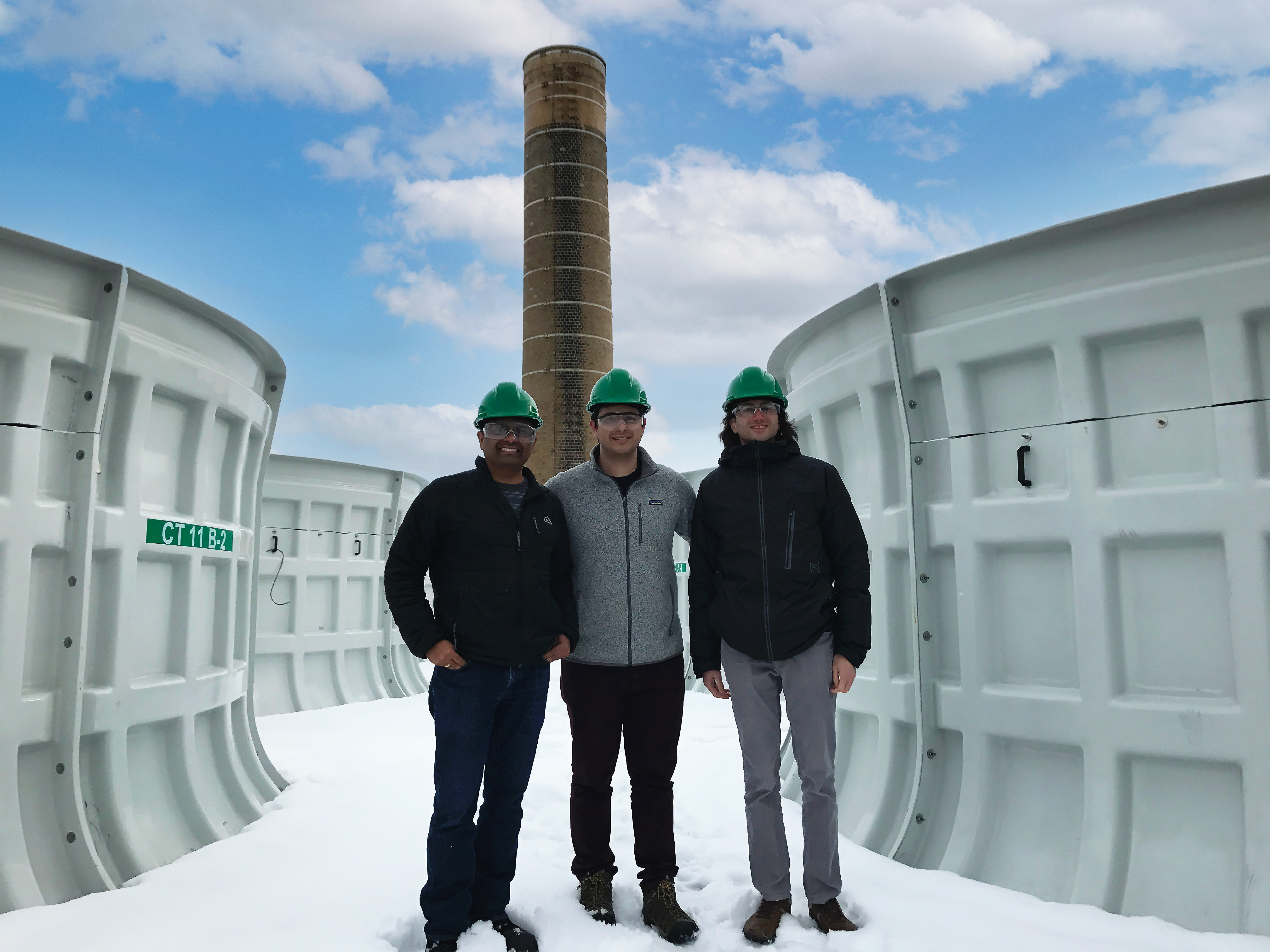 Three people in hard hats stand in the snow in front of a power plant