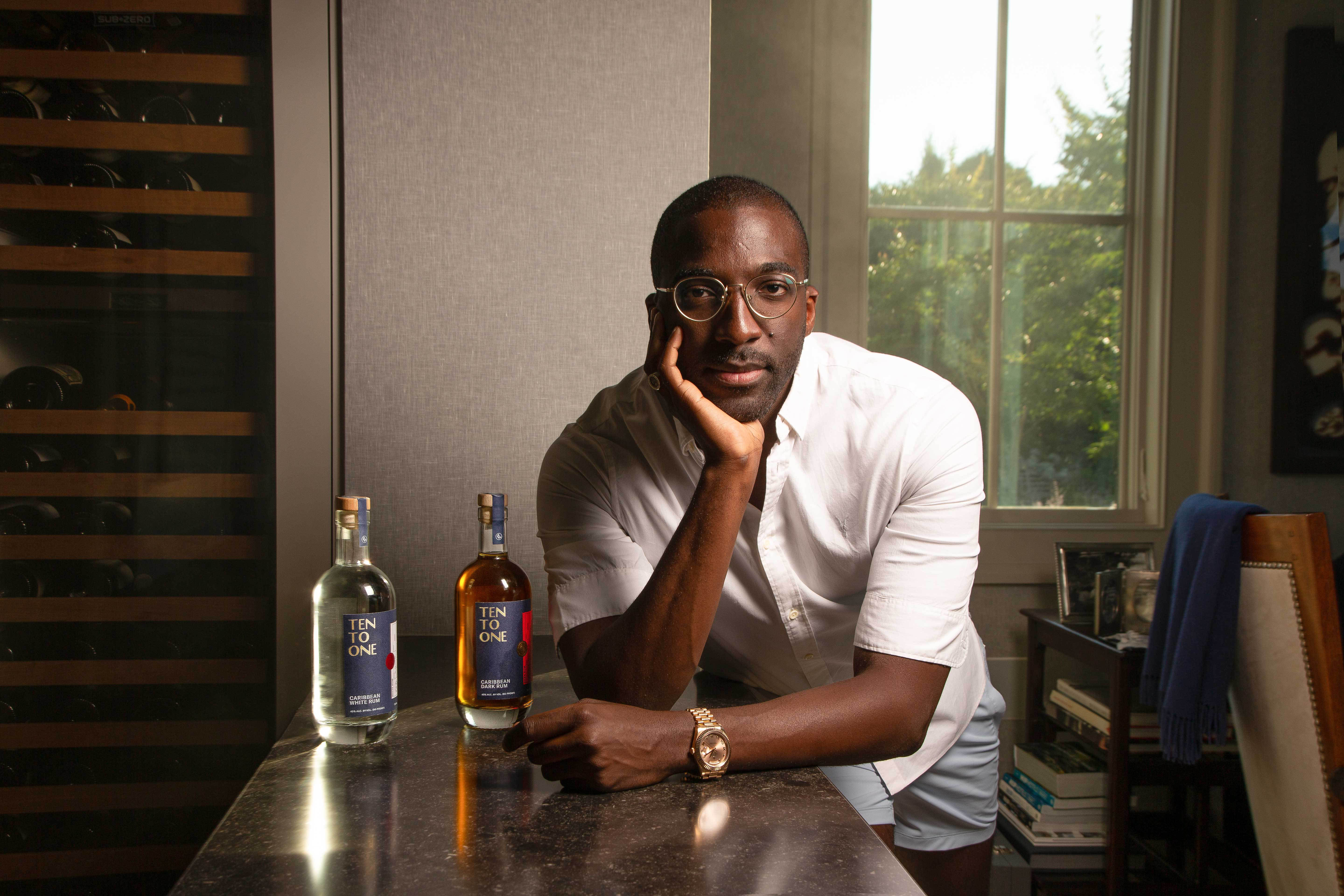 Photo of Marc-Kwesi Farrell leaning on a counter with two bottles of rum to the left a window behind him on the right and a wine fridge on the left