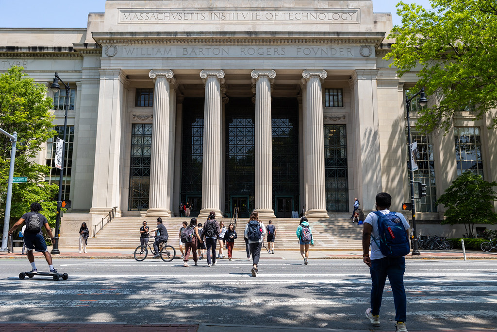 A photo of 77 Mass Ave building at MIT, buildings with columns and students crossing the street in front of the building