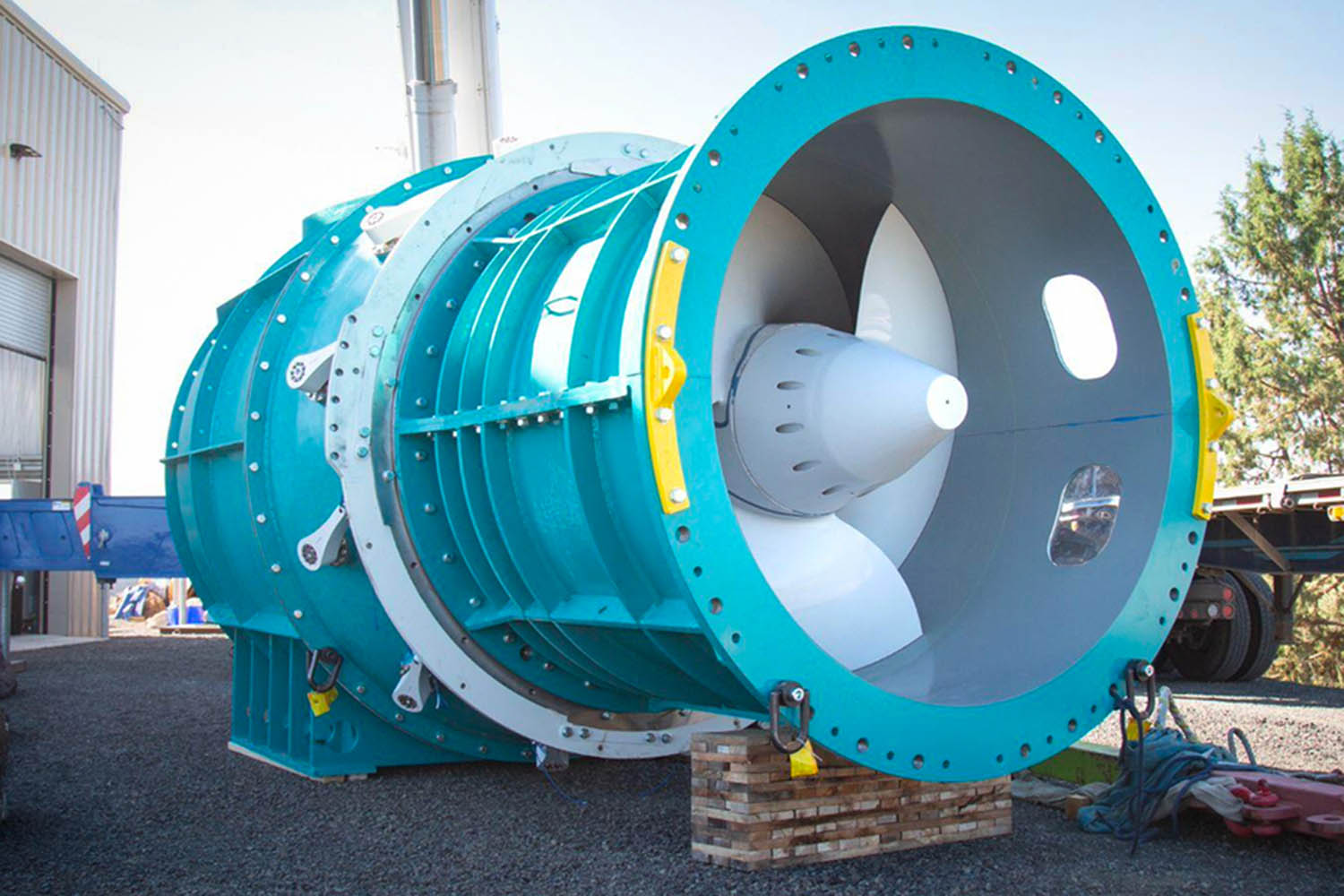 A large turbine is shown in what appears to be a parking lot. The housing is light blue, and the turbine is white. A flatbed truck just visible in the background provides a sense of scale. The turbine is many times taller than the truck.