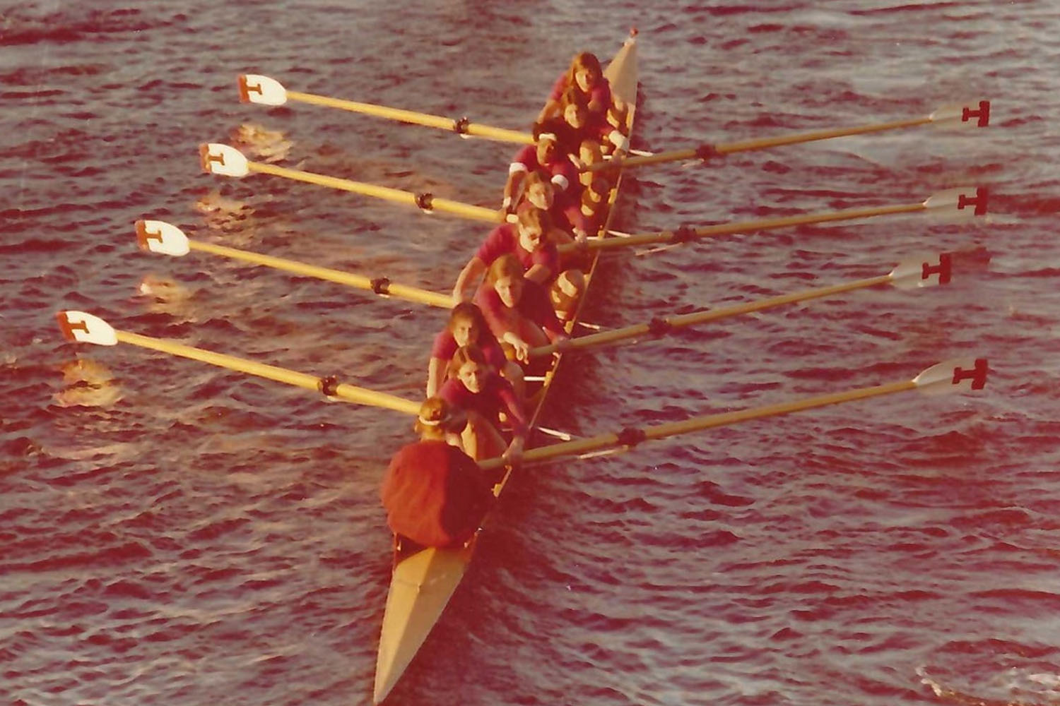An 8-person shell on the water is shown from above with 8 women rowers and a cox