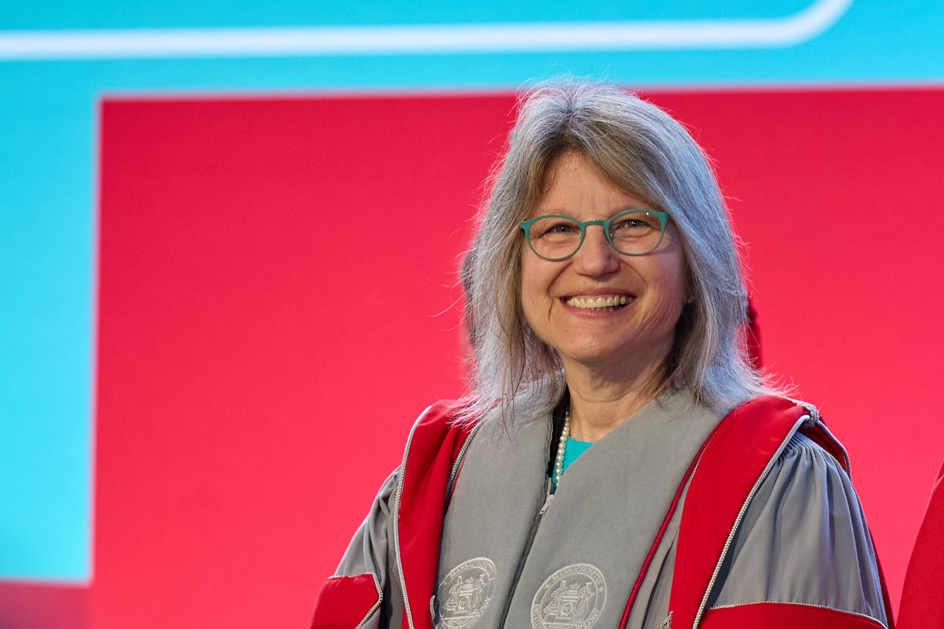 MIT President Sally Kornbluth smiling, in MIT robes, from chest up with red and blue background.