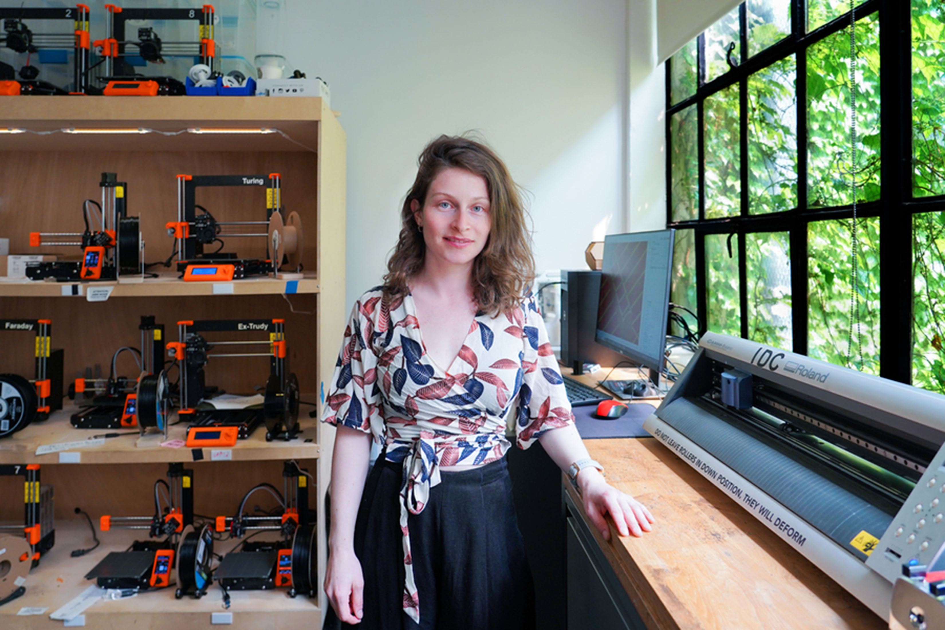 Ganit Goldstein stands in one of MIT's makerspaces. 3D printers are visible in the background. A wall of windows shows greenery outside.
