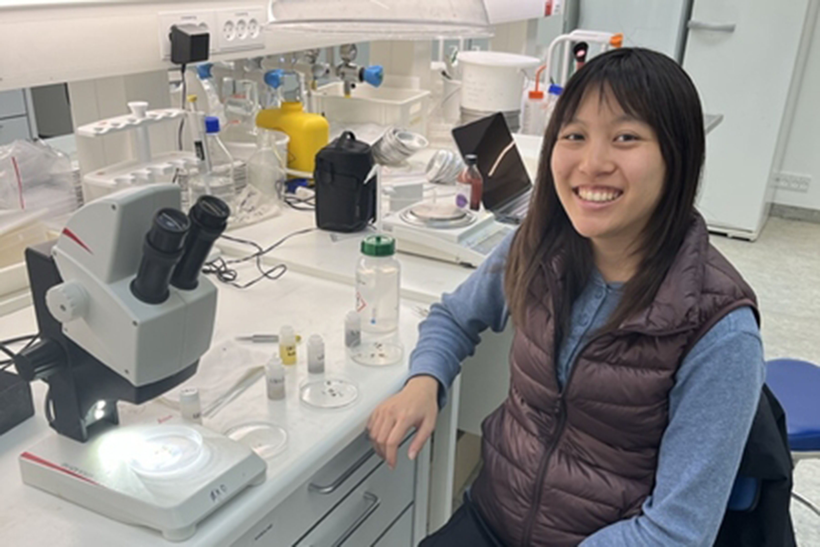 Andrea Lo sits in a lab with a microscope and other lab equipment visible on the counter.