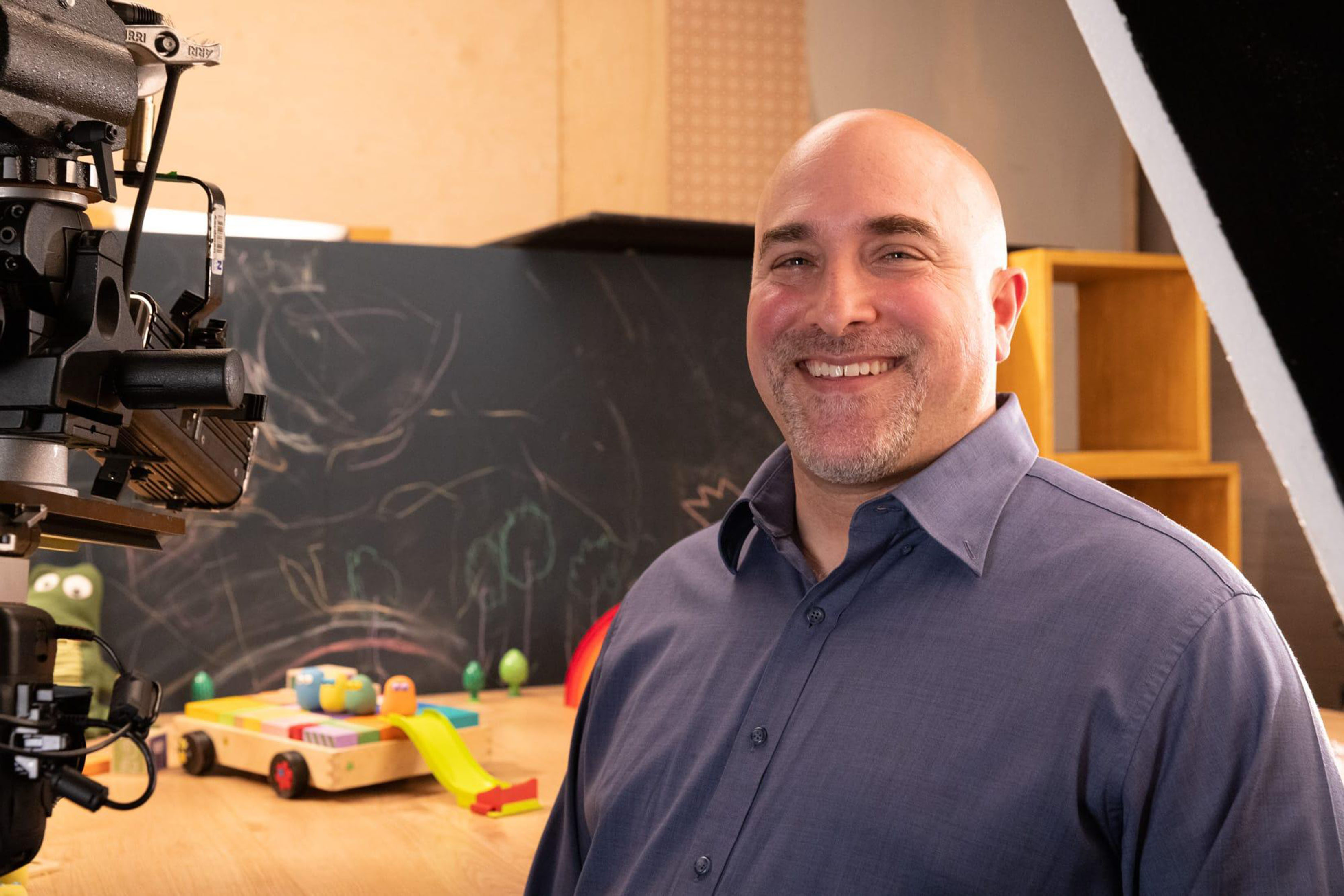 Steven Shapiro is shown beside a table with toys and a blackboard. A movie camera is visible at left.