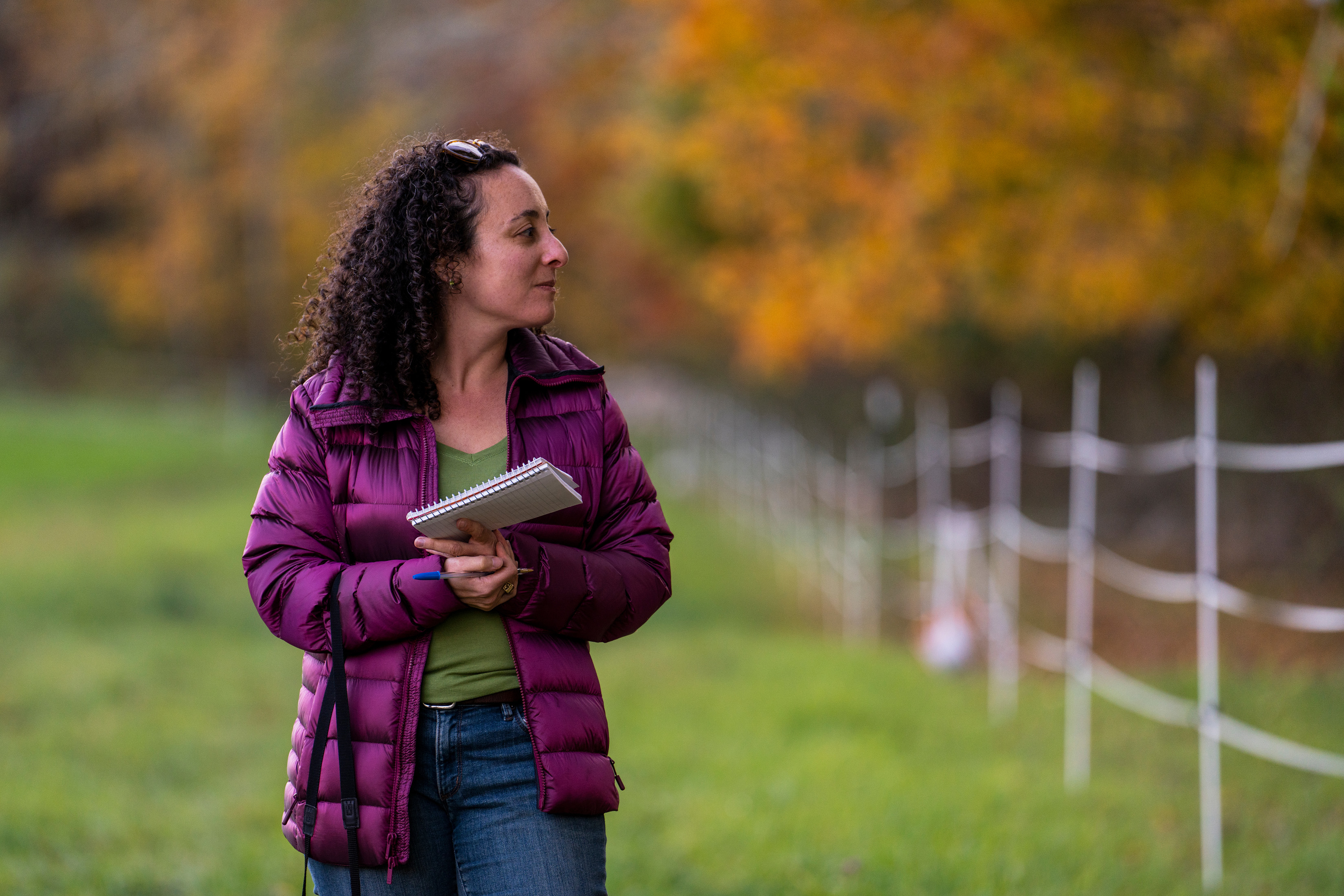Julia Kurnik is shown walking in a field holding a notebook. Fencing and fall leaves can be seen in the blurry background.