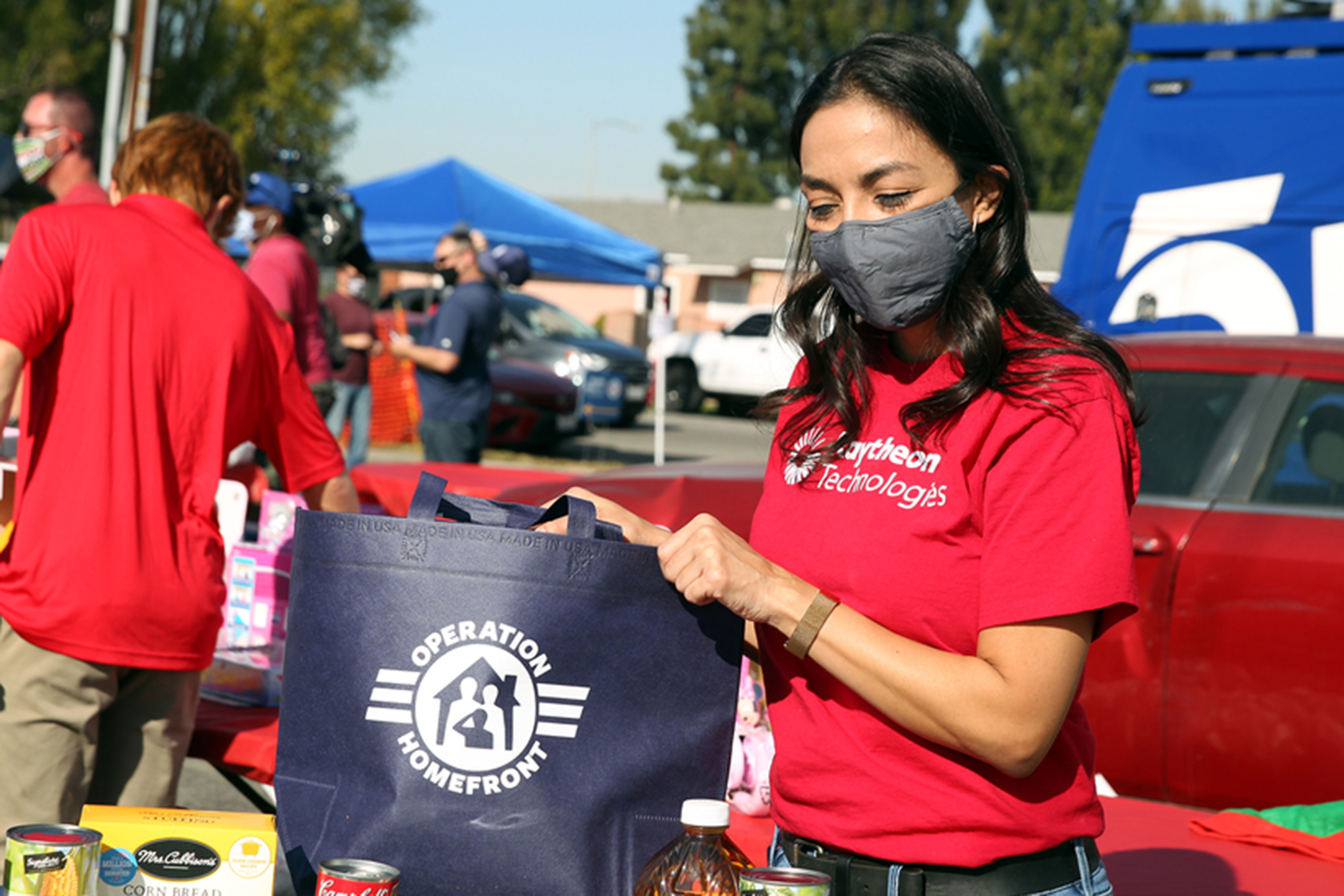 Annabel Flores is shown outside wearing a red Raytheon shirt and gray facemask. She is holding a blue bag that sits on a table with some canned goods.