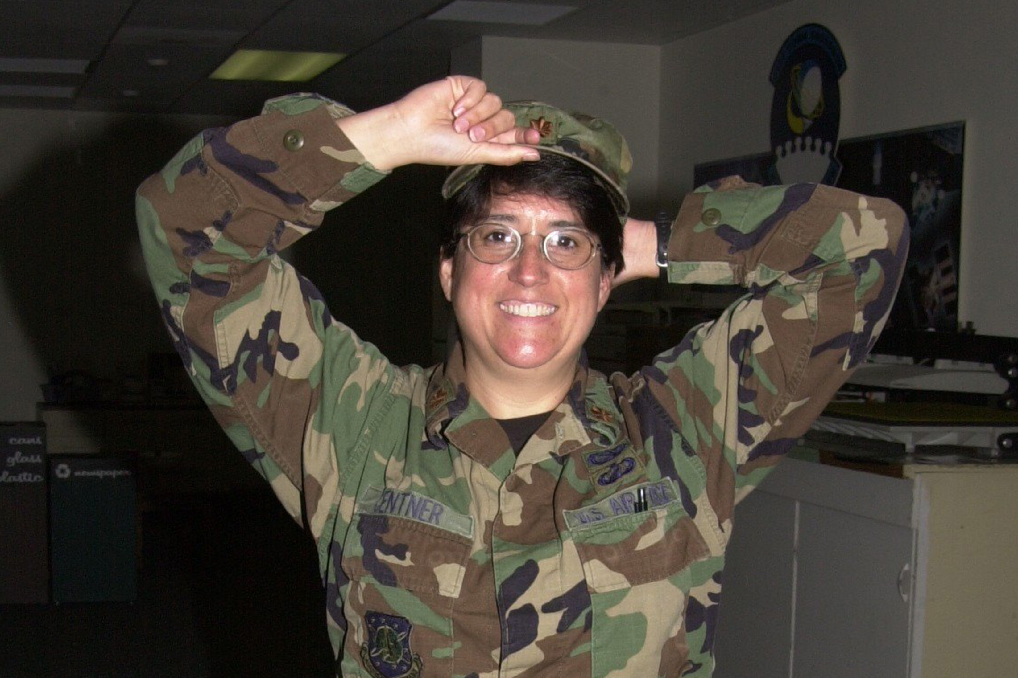 Teri Centner is show from the waist up wearing military fatigues and adjusting her hat with both hands. She is smiling broadly.