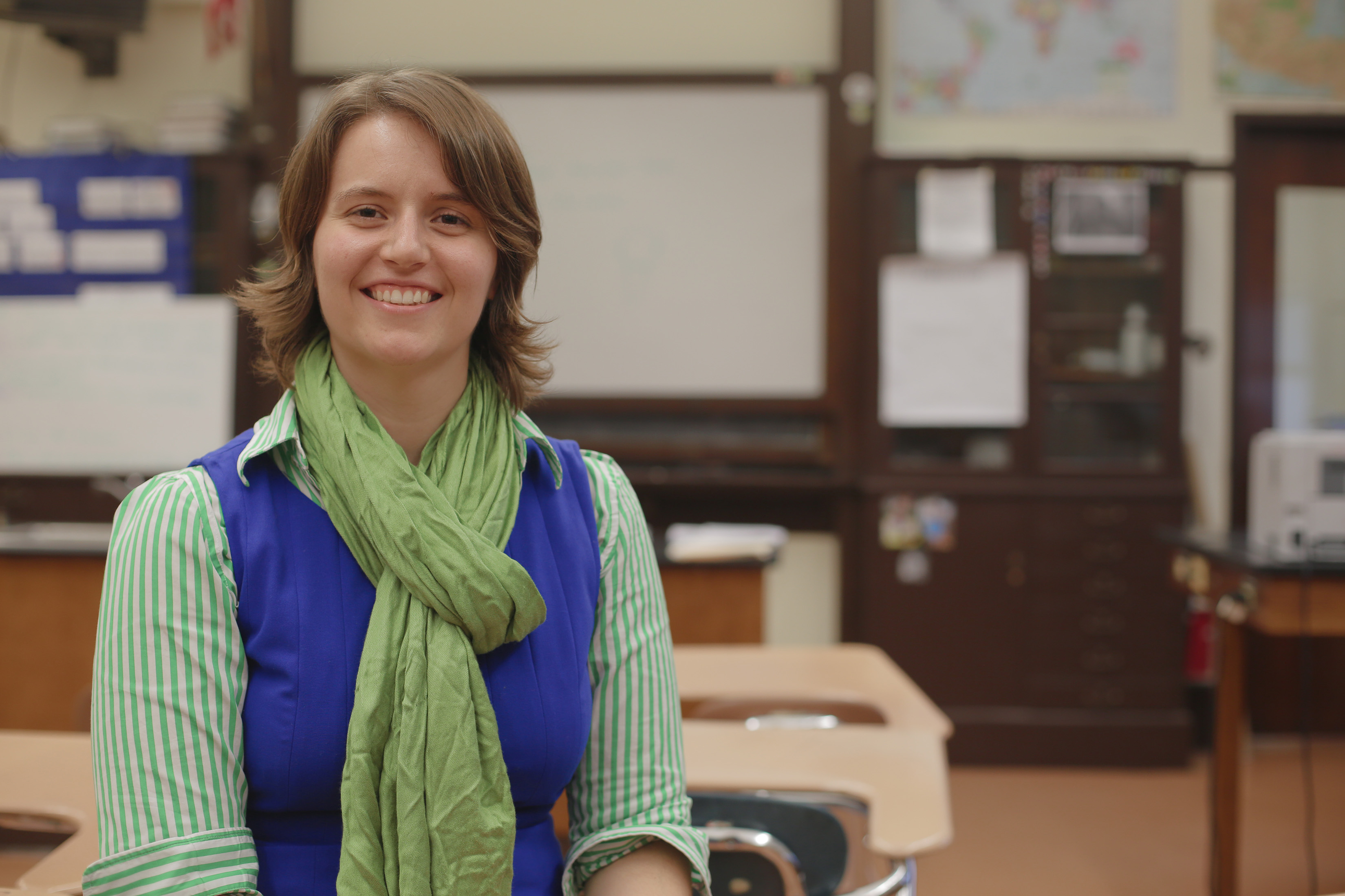 Shannon Morey portrait shows her in a classroom, which is blurry in the background. She wears a green scarf, blue vest, and striped green collared shirt.