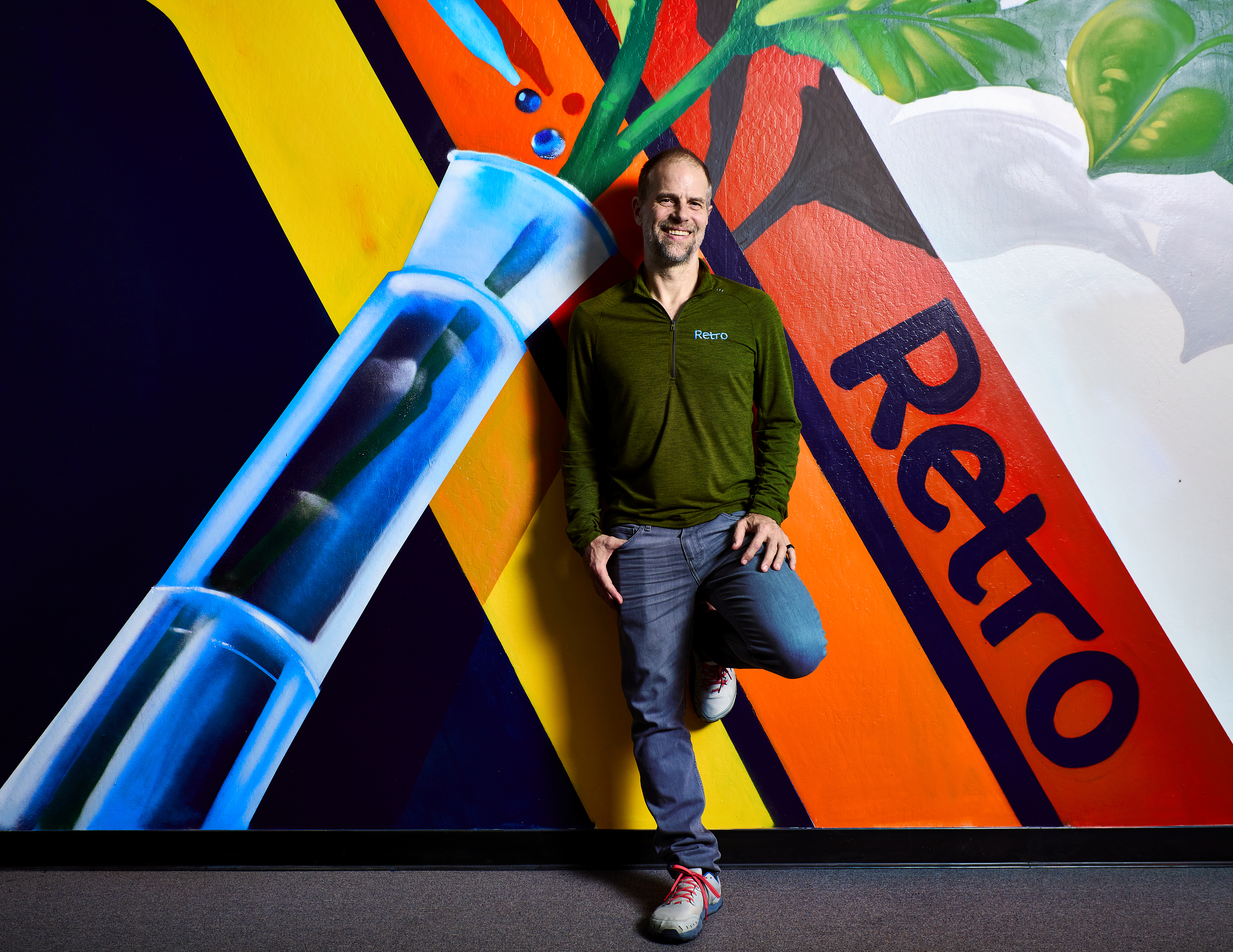 Joe Betts-LaCroix leans against a wall with a mural on it, featuring a test tube with a green stem coming out the top and yellow orange and red lines, with the word Retro written on the red line