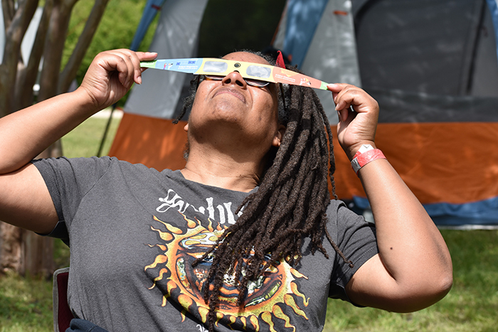 A photo of MIT alum Dara Norman with both hands holding a strip of material over her eyes while gazing up into the sky, with grass and a tent behind her