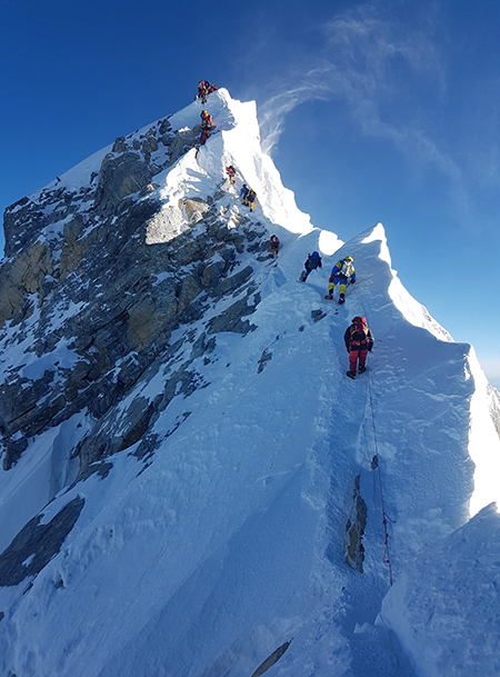 A photo of a line of people climbing up the face of Mount Everest with snow on the mountain and blue skies behind