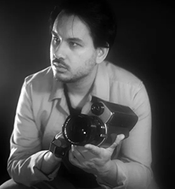 A black and white portrait of Suneil Sanzgiri holding a large camera. The frame shows him from the elbows up and he is looking off to one side.
