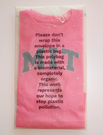 A pink MIT shirt appears wrapped in see-through packaging. The packaging reads: Please don't wrap this envelope in a plastic bag. This polybag is made with a biomaterial, completely organic. This work represents our hope to stop plastic pollution.