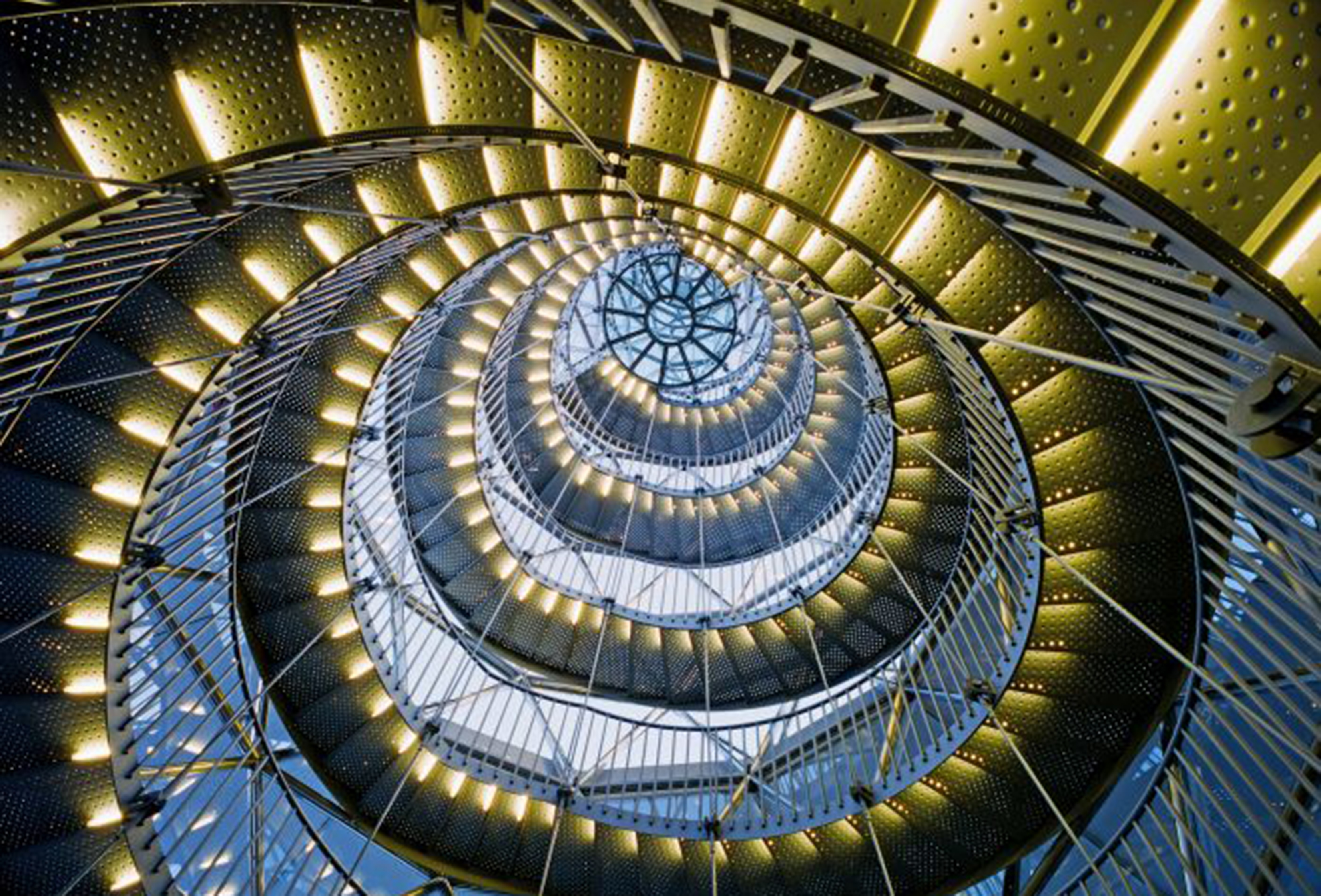A structure of gold colored metal stairs and interlacing framework spirals upward toward a skylight.