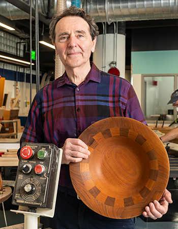 Irving Fischman faces the camera holding an intricately inlaid wooden bowl. In the foreground, there is a panel of buttons for a machine. Just visible in the background is another person working in the woodshop.