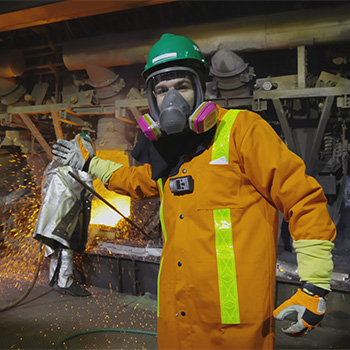 James Dingley wears a protective orange suit, hard hat, and breathing apparatus in an industrial setting. He is gesturing toward a person in the background who is dressed in silver protective gear and is leaning toward a bright orange furnace. Sparks can be seen on the floor.