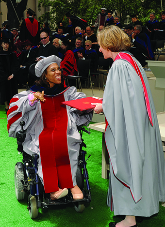 MIT President Hockfield, at right, wearing gray MIT robes, presents a red-cased diploma to Allison V. Thompkins, who wears gray MIT PhD robes and is seated in a wheelchair. They are outside and some audience members are visible in the background. 