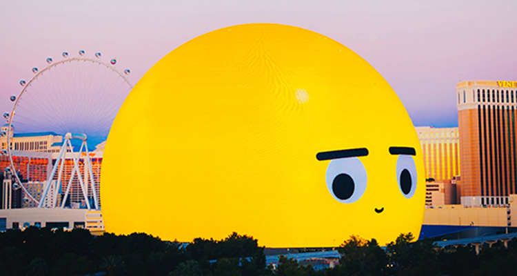 A giant yellow sphere dominated the Las Vegas skyline. On the right side, it has the face of an emoji. A Ferris wheel is to the left and other buildings to the right in the background.