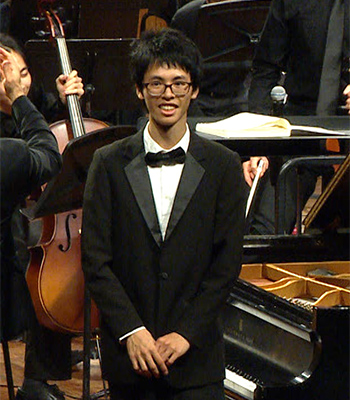 Holden Mui stands with hands clasped in front. He is wearing a tuxedo. In the background, parts of a piano, cello and other orchestra elements can be seen.